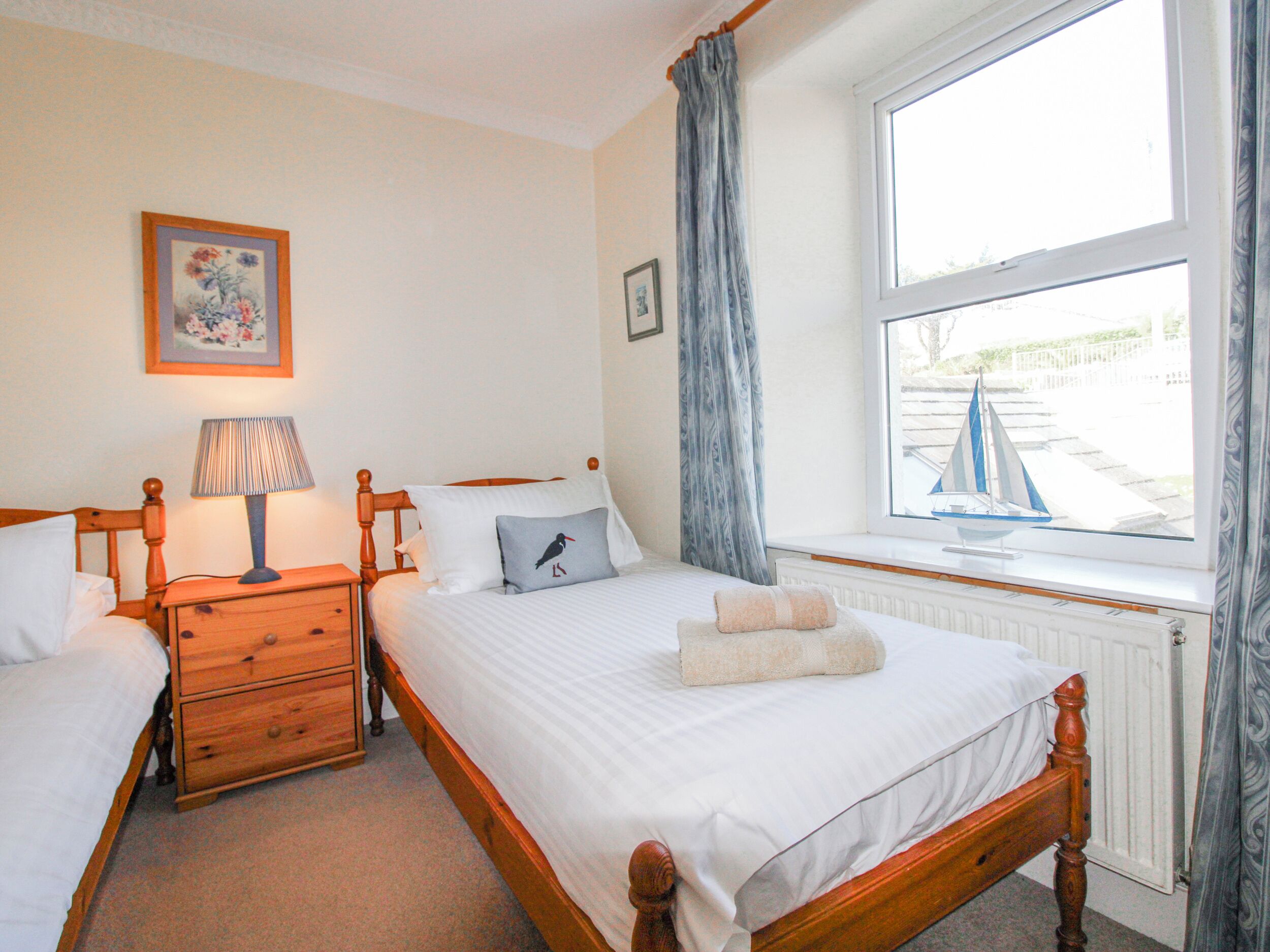 A bedroom with a wooden bed, a nightstand, a lamp, a window with a view of a sailboat, and a framed picture on the wall.