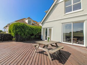 Eine Holzterrasse mit Picknicktisch, einer Glastür und Blick auf ein Haus mit weißer Fassade und einer grünen Hecke.