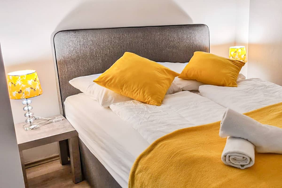 A neatly made bed with white linens, two mustard yellow pillows, and rolled towels on a mustard yellow blanket.