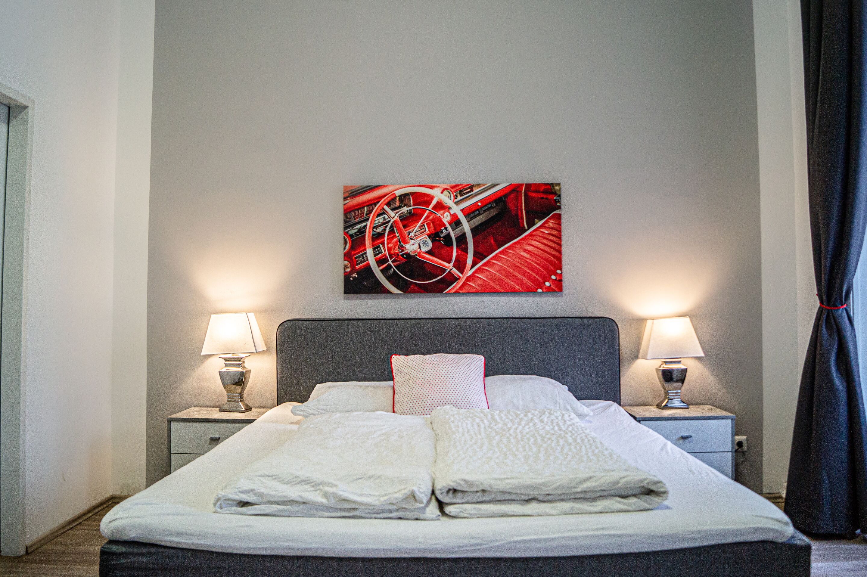 Una cama bien hecha con sábanas blancas, dos lámparas de noche y un cuadro abstracto rojo en la pared.