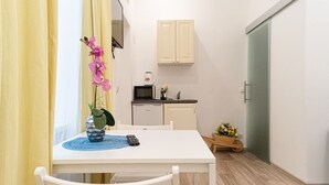 Una habitación pequeña y limpia, con una mesa blanca, una silla, un control remoto, una planta pequeña en maceta y una zona de cocina con armarios y electrodomésticos.