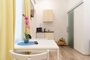A small, clean room with a white table, a chair, a remote control, a small potted plant, and a kitchen area with cabinets and appliances. - Mariahilfer Boutique Rooms - Virtual Reception & Self-Checkin (Vienna)