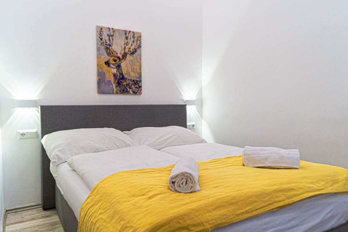 A neatly made bed with a white and yellow bedspread, a rolled towel on the bed, and a painting on the wall.