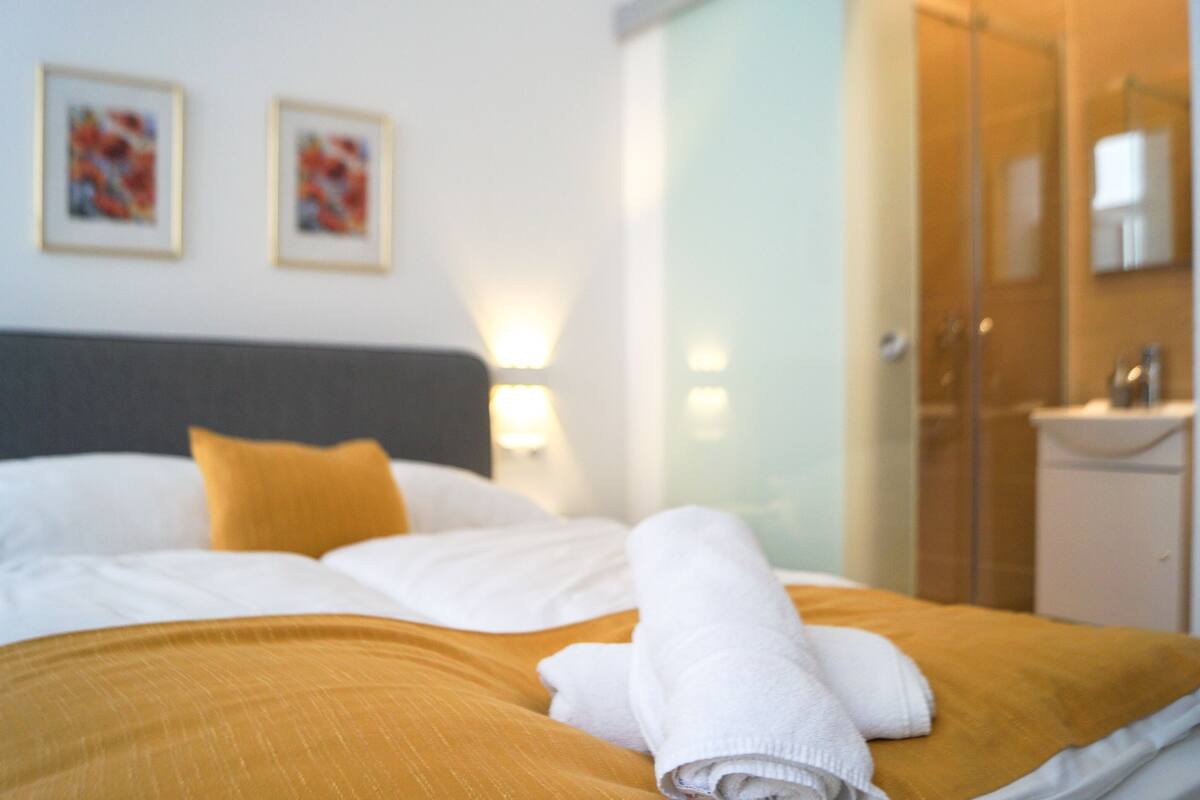 A neatly made bed with white and mustard bedding, a folded towel, and two framed artworks on the wall.