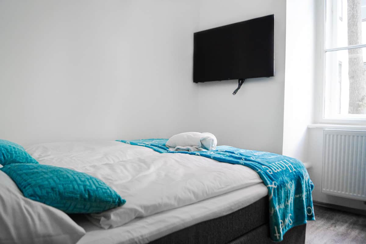A bedroom with a bed, a flat-screen TV mounted on the wall, and a window with a view of trees.