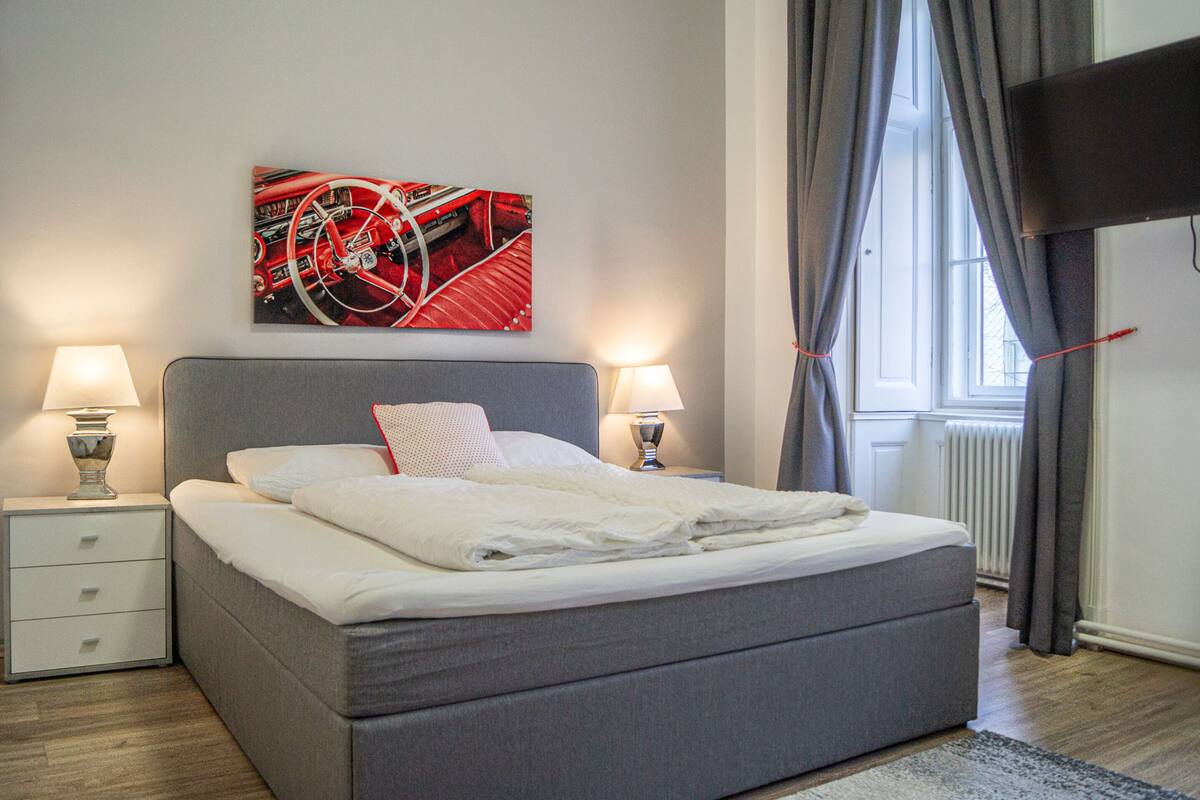 A neatly made bed with white linens, two bedside tables with lamps, a red abstract painting on the wall, and a window with curtains.