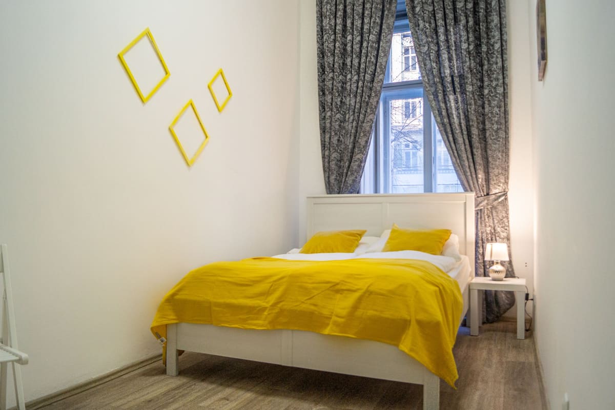 A bedroom with a bed, a nightstand, a window with curtains, and wall-mounted picture frames.