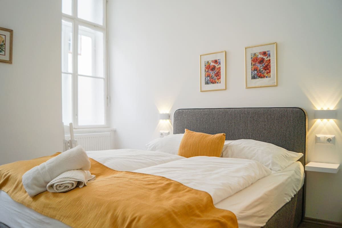 A neatly made bed with a white and mustard-colored bedspread, two framed floral artworks on the wall, and bedside lamps.