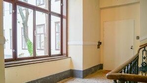 Un pasillo con una escalera de madera, una puerta blanca y un ventanal con vistas a un árbol y edificios.
