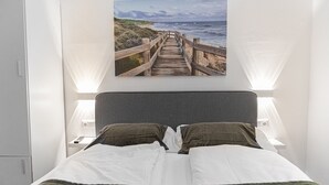 Un dormitorio con una cama, dos lámparas de noche, una mesita de noche, un cuadro en la pared de una playa y un armario.
