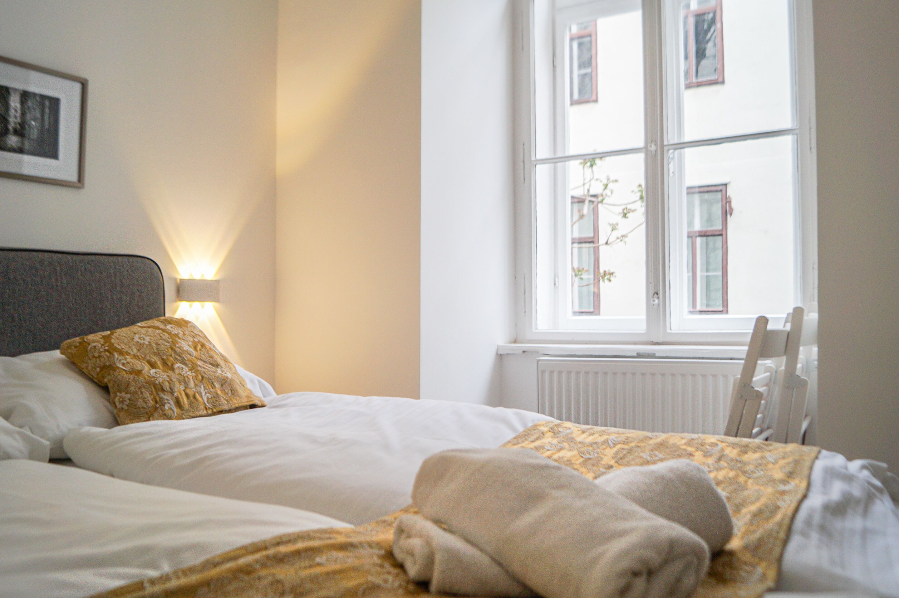 Un lit bien fait, agrémenté d’un coussin à motifs, une lampe de chevet et une vue sur un bâtiment par la fenêtre.