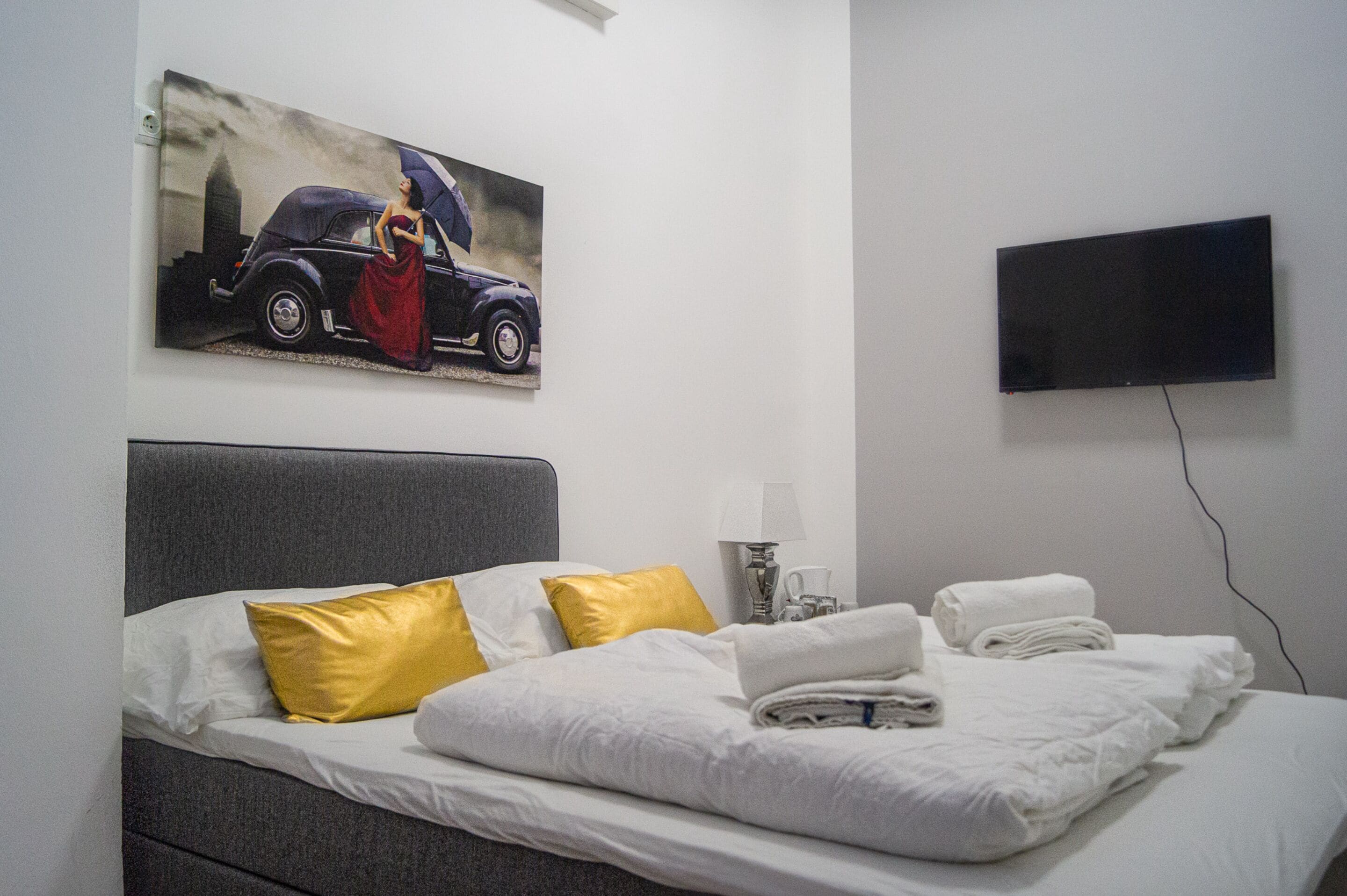 Habitación de hotel con una cama, un cuadro de un automóvil y una mujer, un televisor de pantalla plana y una mesita de noche con una lámpara y un vaso.