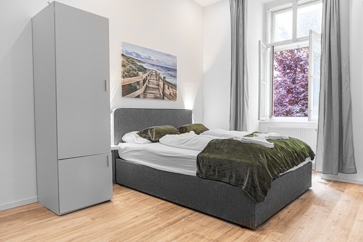 A bedroom with a bed, a grey wardrobe, a window with curtains, and a wall-mounted picture.