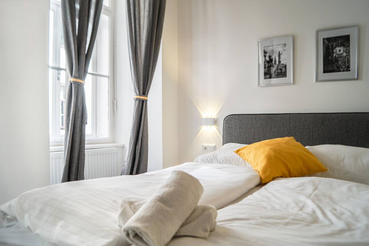 A neatly made bed with white linens and a single yellow pillow, a bedside lamp, and two framed pictures on the wall.