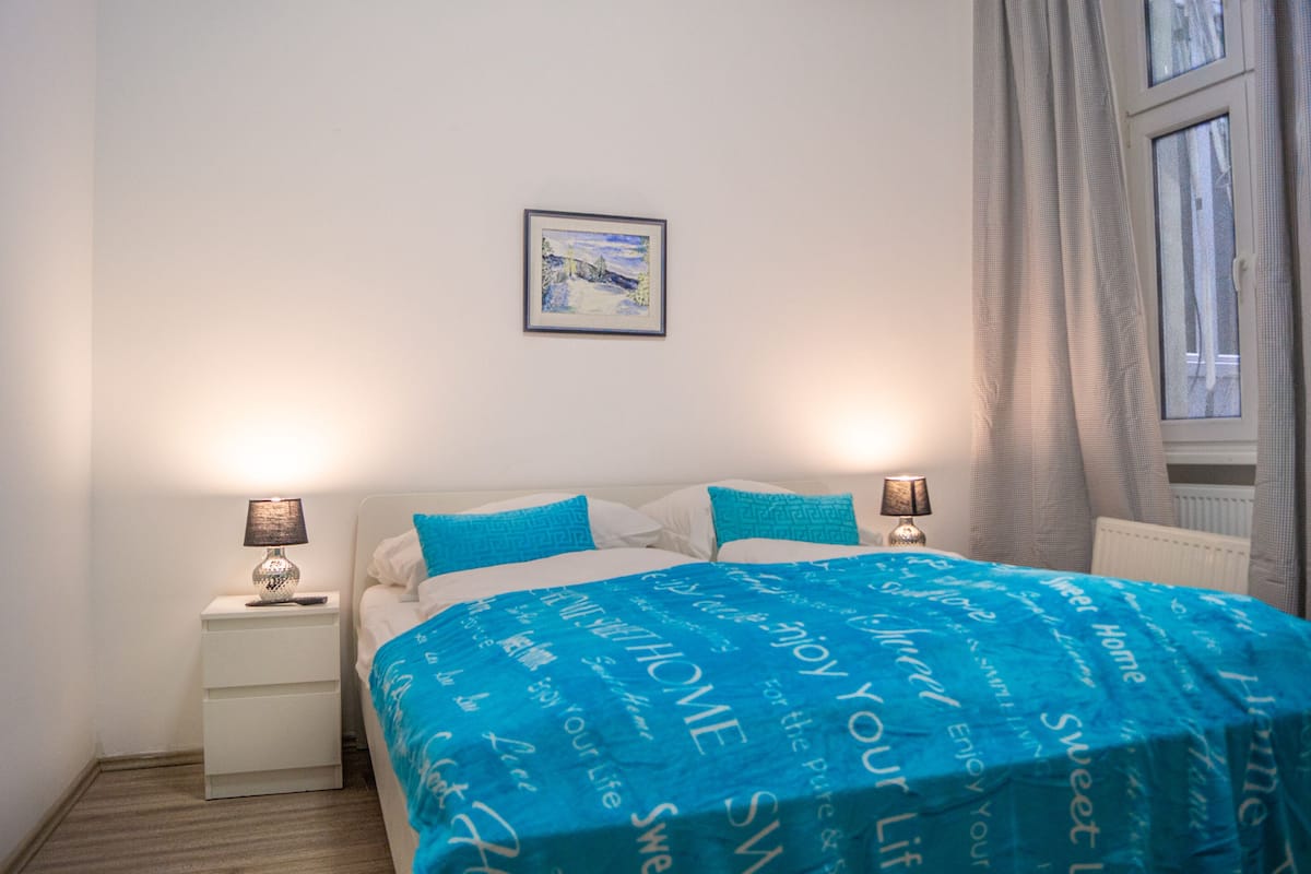 A bedroom with a bed, two bedside lamps, a nightstand, a framed picture on the wall, and a window with curtains.