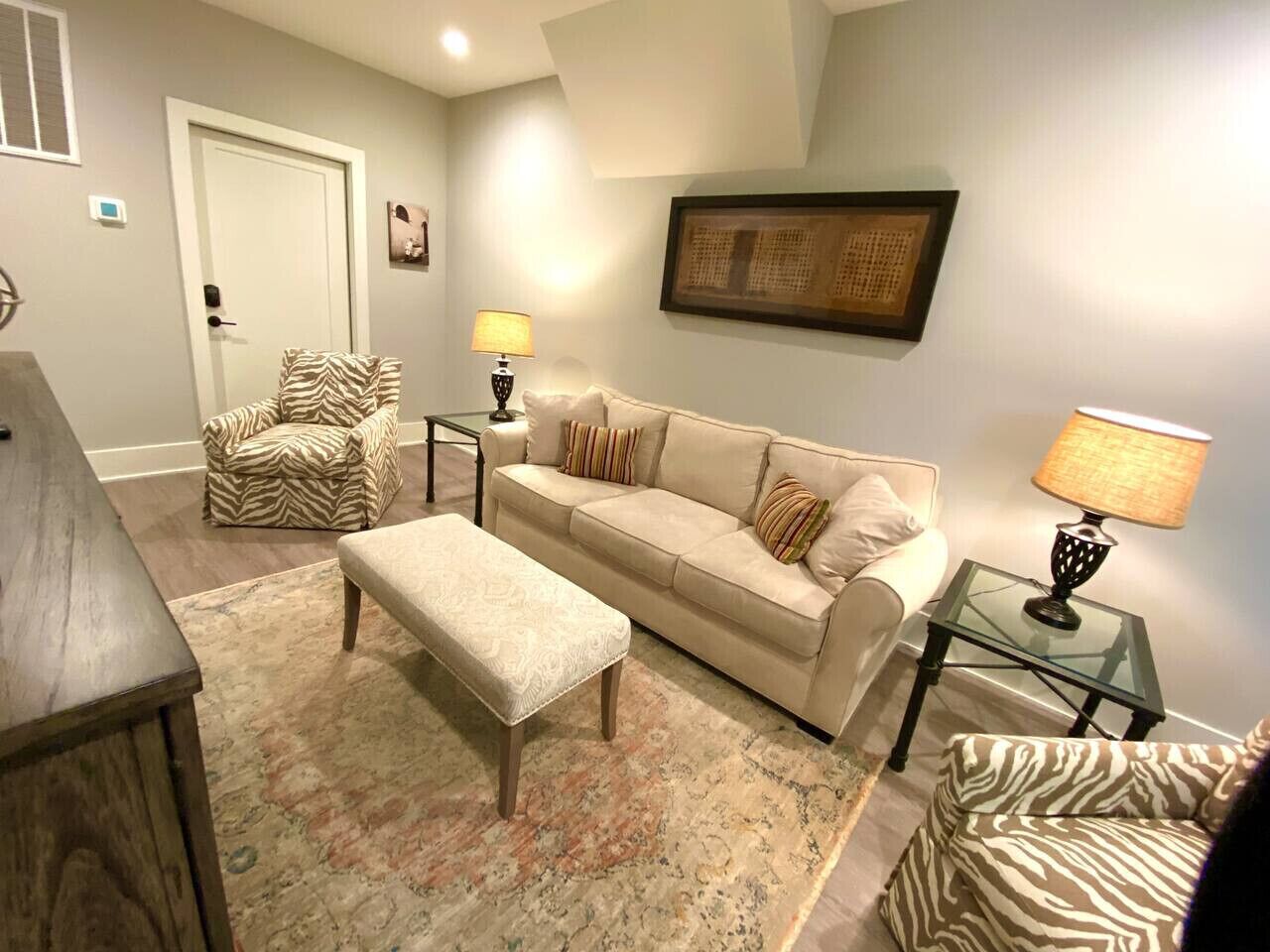 Un salon avec un canapé beige, un pouf, un fauteuil à motifs zébrés, une table basse en verre et une lampe.