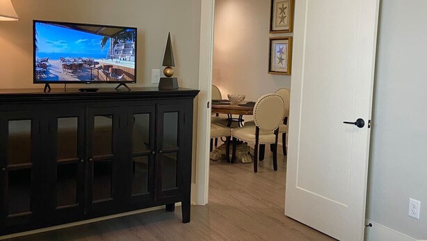 Una habitación con un armario de madera oscura, un televisor mostrando una escena de playa, una zona de comedor con mesa y sillas, y una puerta blanca.