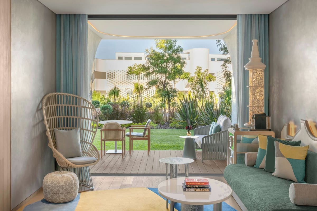 A modern hotel room with a view of a lush garden, a wicker chair, a sofa, a small table, and a decorative lamp.