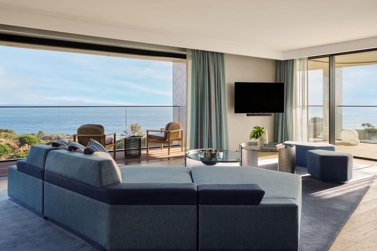 A modern living room with a large sofa, a coffee table, and a flat-screen TV. There is a balcony with a view of the ocean.