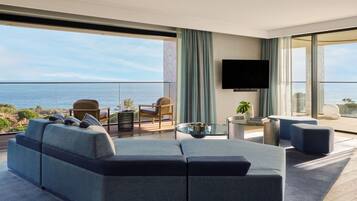 A modern living room with a large sofa, a coffee table, and a flat-screen TV. There is a balcony with a view of the ocean.