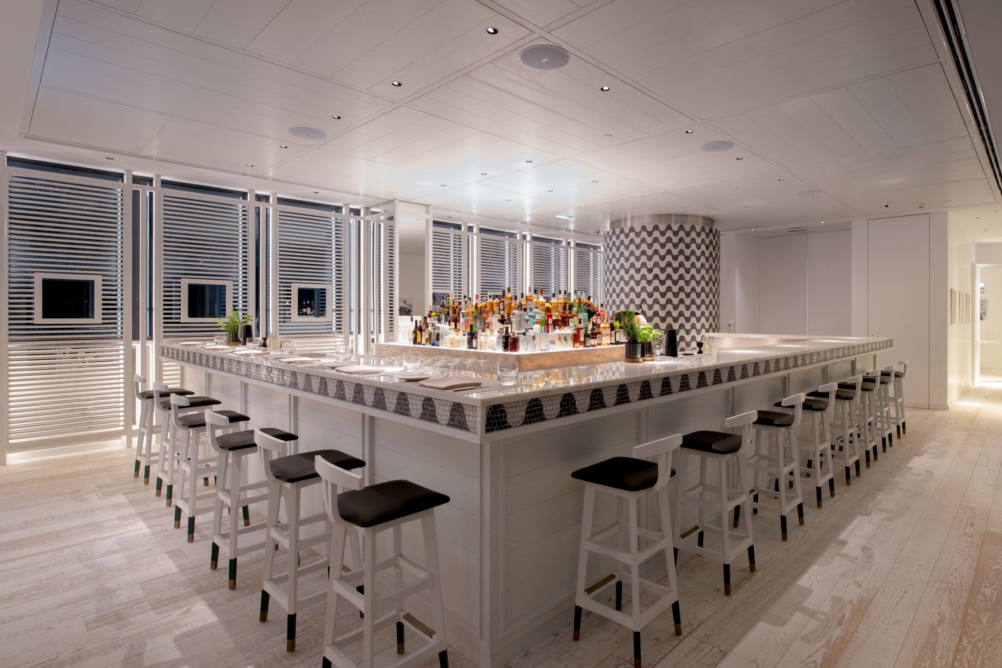 Een moderne bar met een lange toonbank, meerdere krukken en een divers aanbod aan flessen.