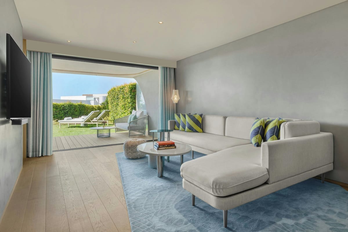 A modern living room with a sofa, coffee table, and a TV. There is a view of a garden and a balcony outside.