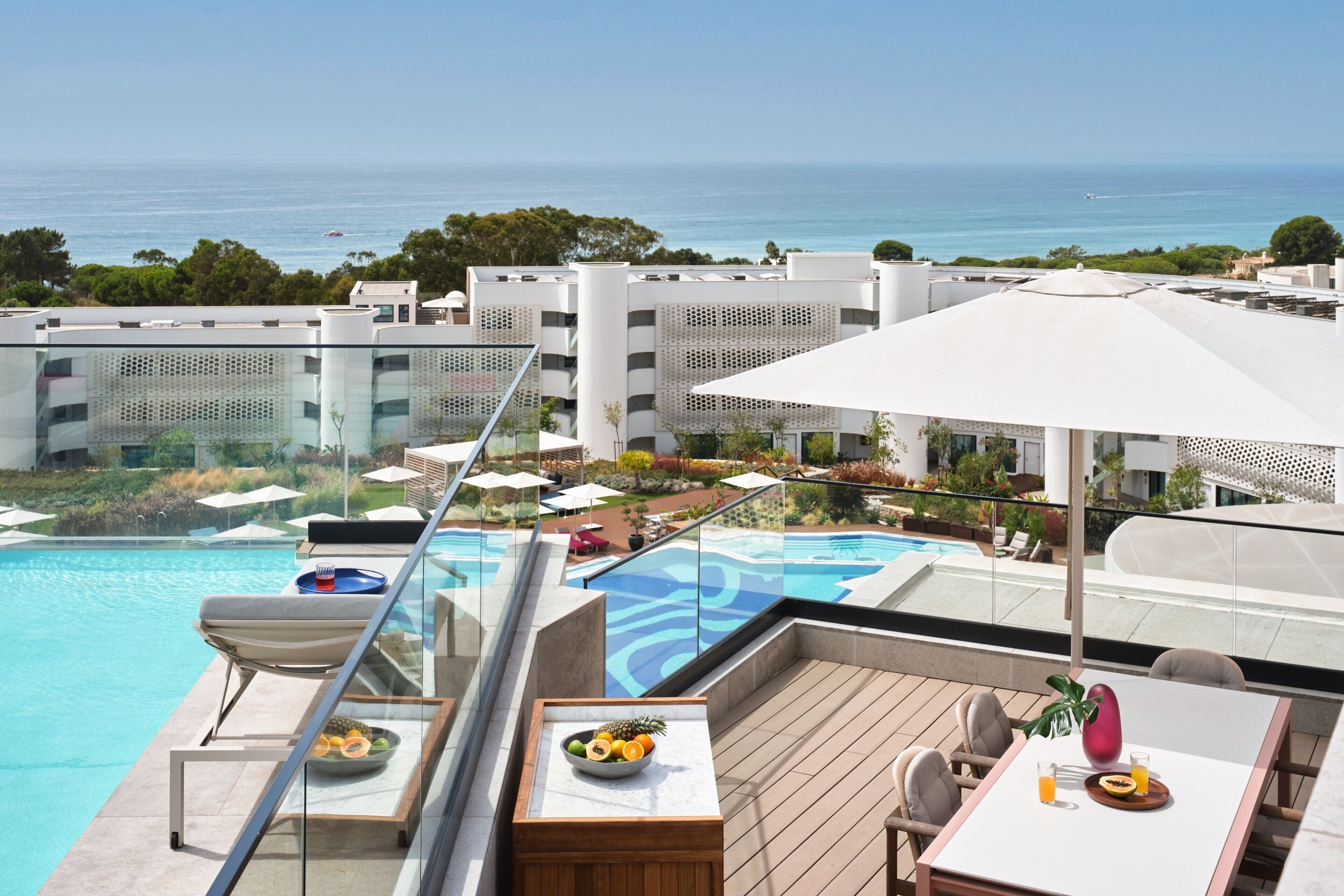 Ein Poolbereich auf dem Dach mit Blick aufs Meer, Liegestühle und ein Esstisch mit Obst und Getränken.