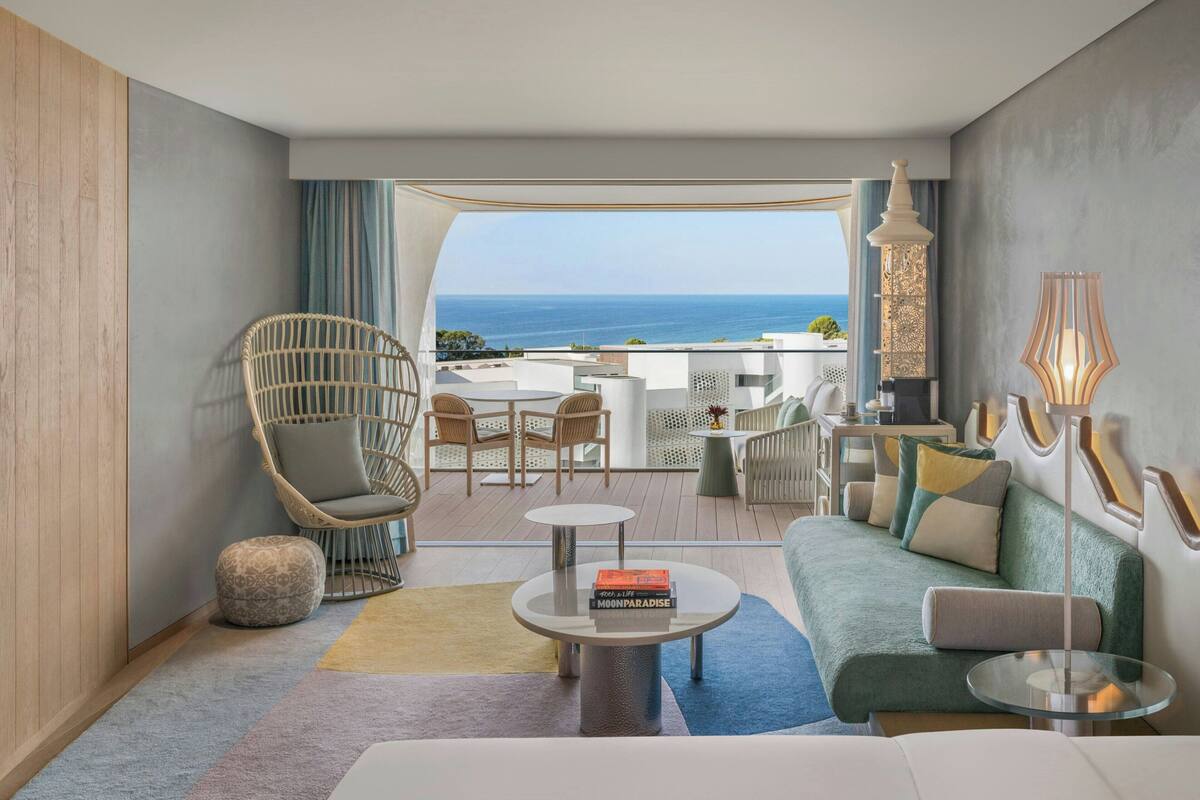 A modern hotel room with a balcony, a wicker chair, a sofa, a coffee table, and a view of the sea.