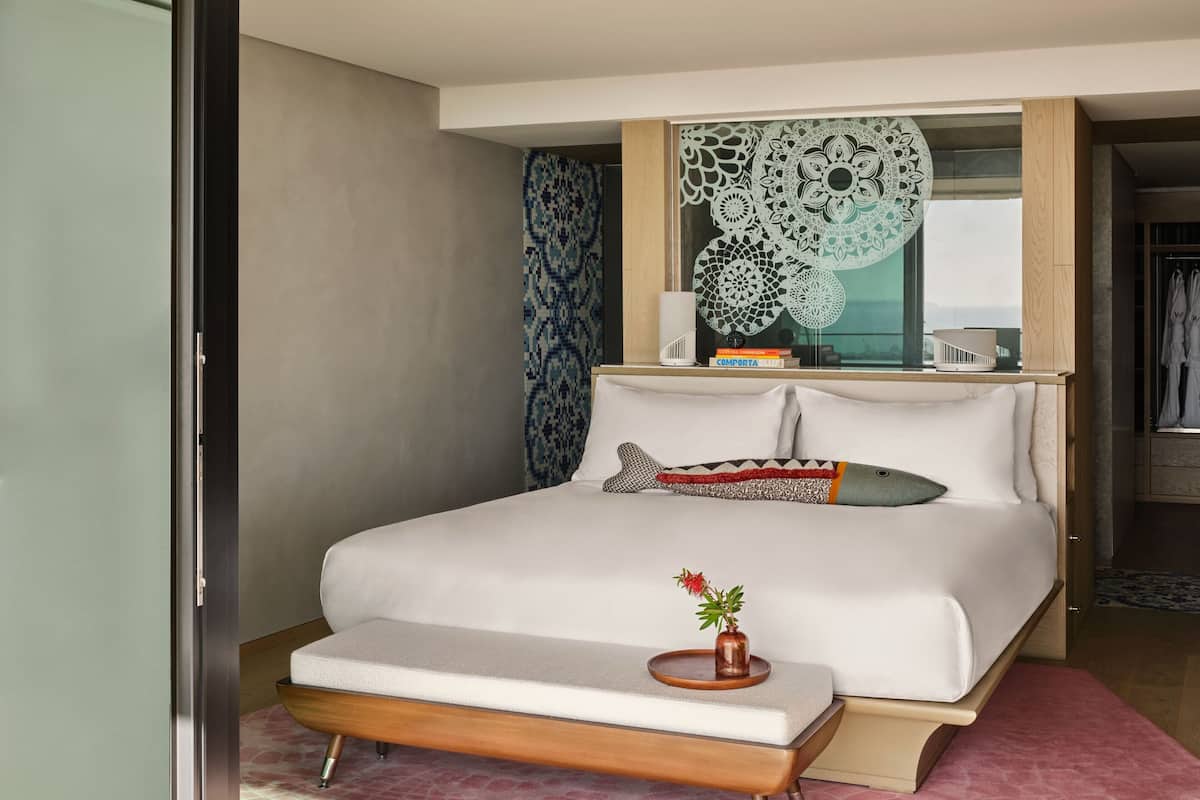 A modern hotel room with a large bed, a bedside table with a lamp, a decorative mirror, and a view of the outdoors.