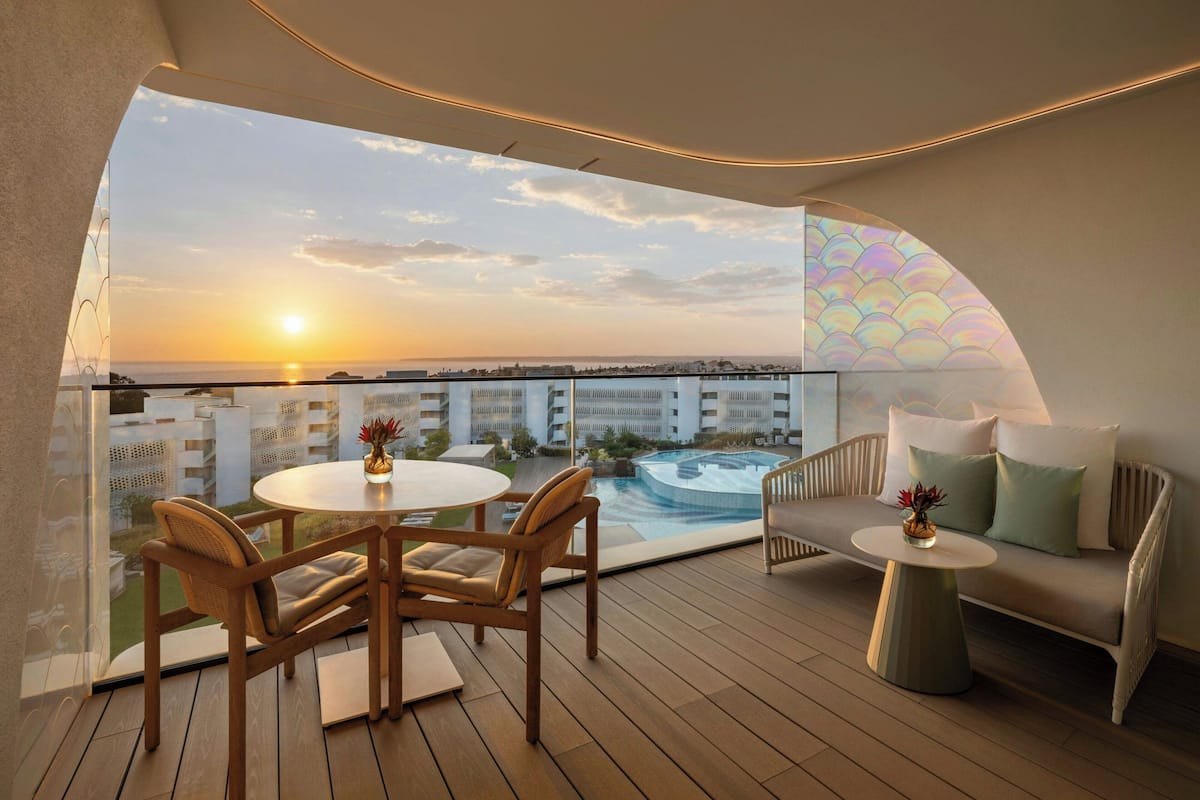 A balcony with a round table and chairs, a sofa, and a view of a swimming pool and buildings during sunset.