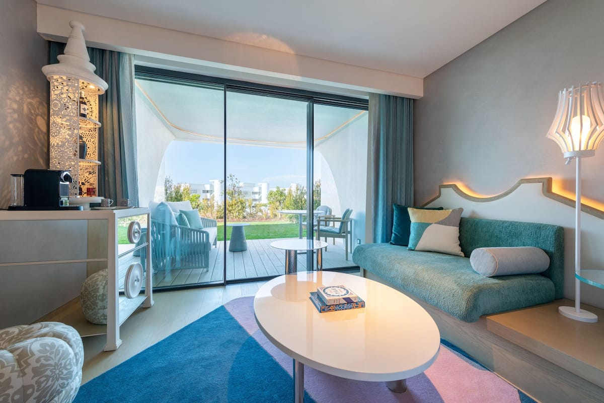 A modern hotel room with a large sliding glass door leading to a balcony, a sofa, a coffee table, and a lamp.