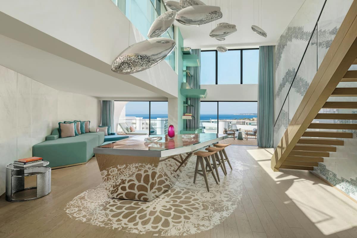 A modern interior with a central island, bar stools, a staircase, and a view of the sea.