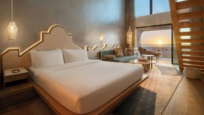 Una habitación de hotel moderna con una cama grande, una zona de estar con sofá y mesa de centro, y vistas al atardecer a través de las ventanas.