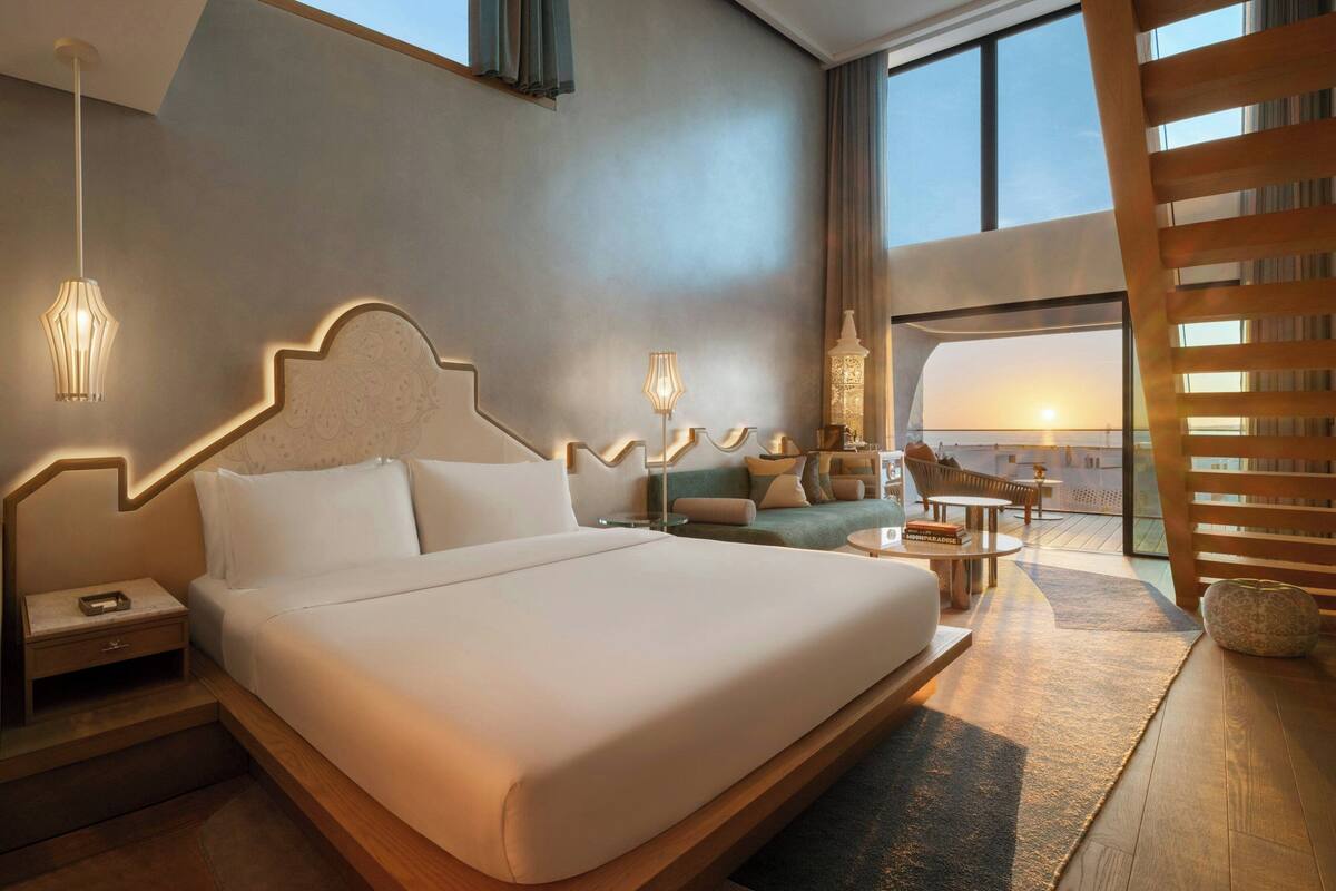 A modern hotel room with a large bed, a sitting area with a sofa and coffee table, and a view of the sunset through the windows.