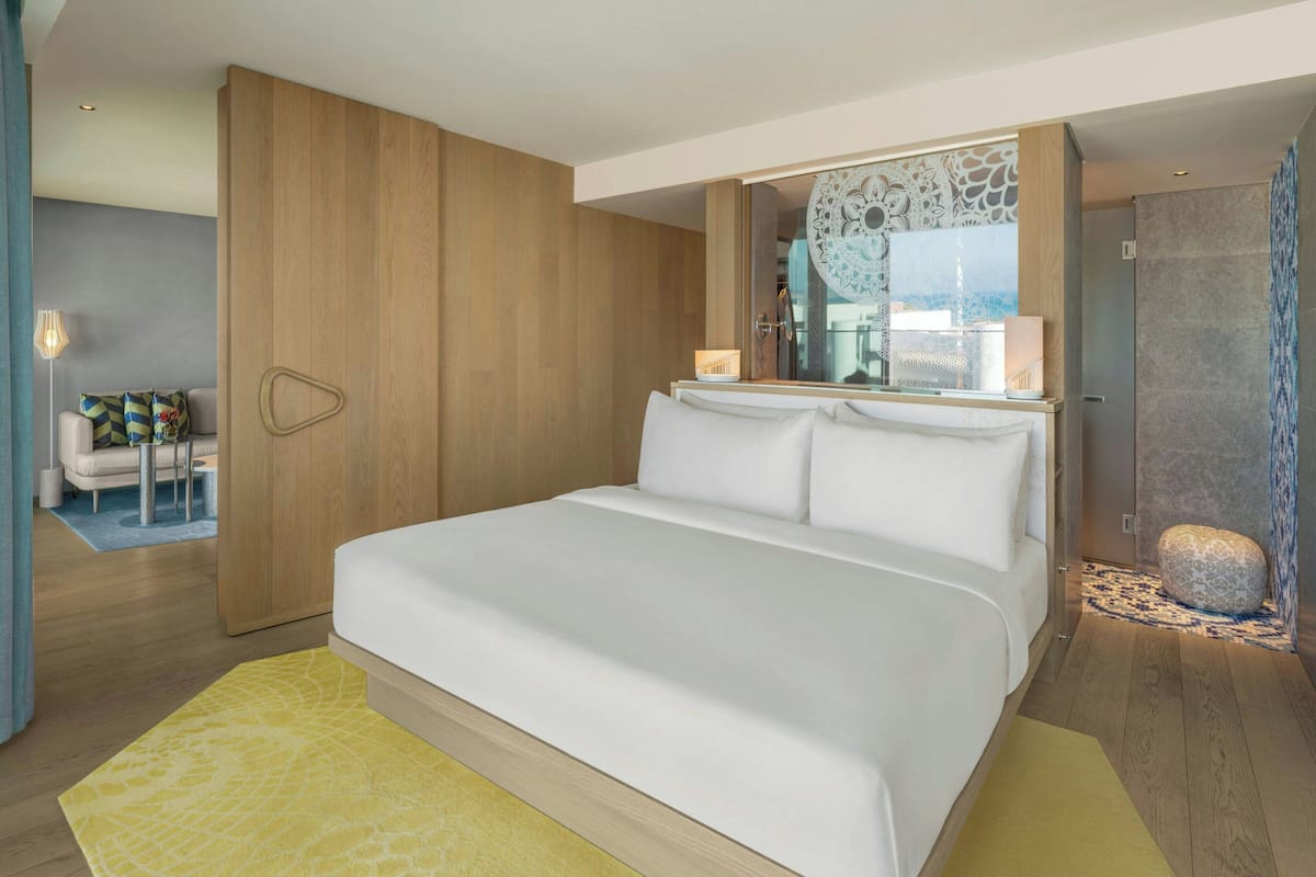 A modern hotel room with a large bed, a wooden wardrobe, a sitting area with a chair and a small table, and a bathroom with a shower and a mirror.