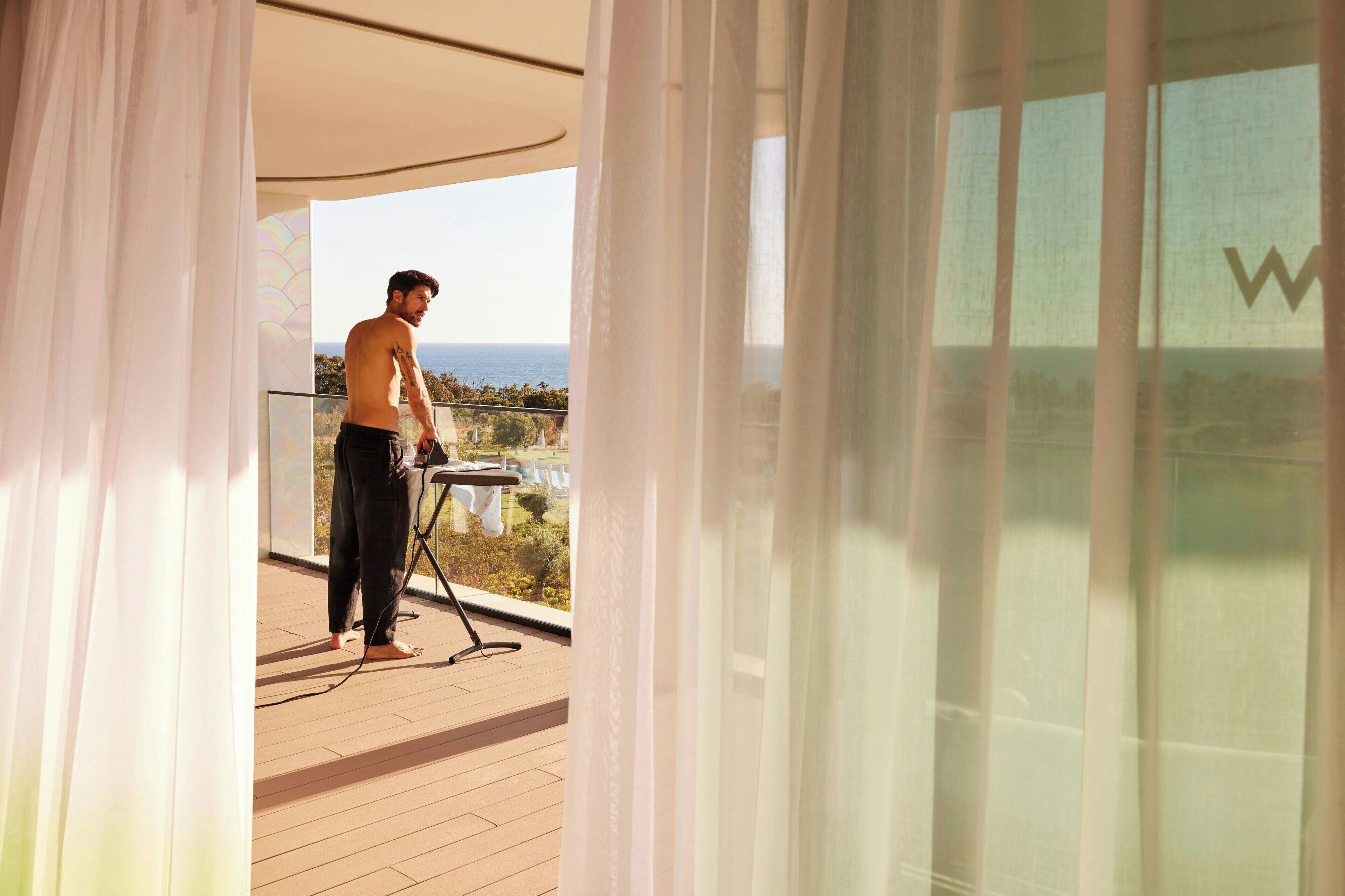 A man ironing on a balcony with a view of the sea.