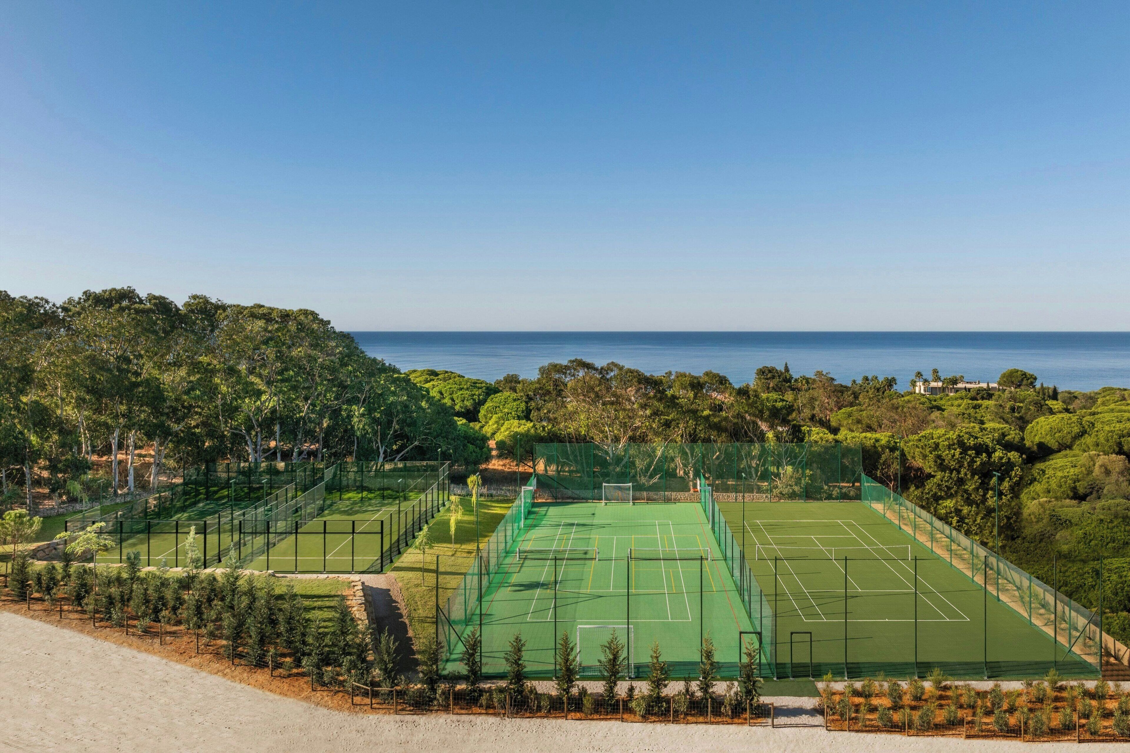 A tennis court surrounded by trees and hills, with a view of the sea in the background.