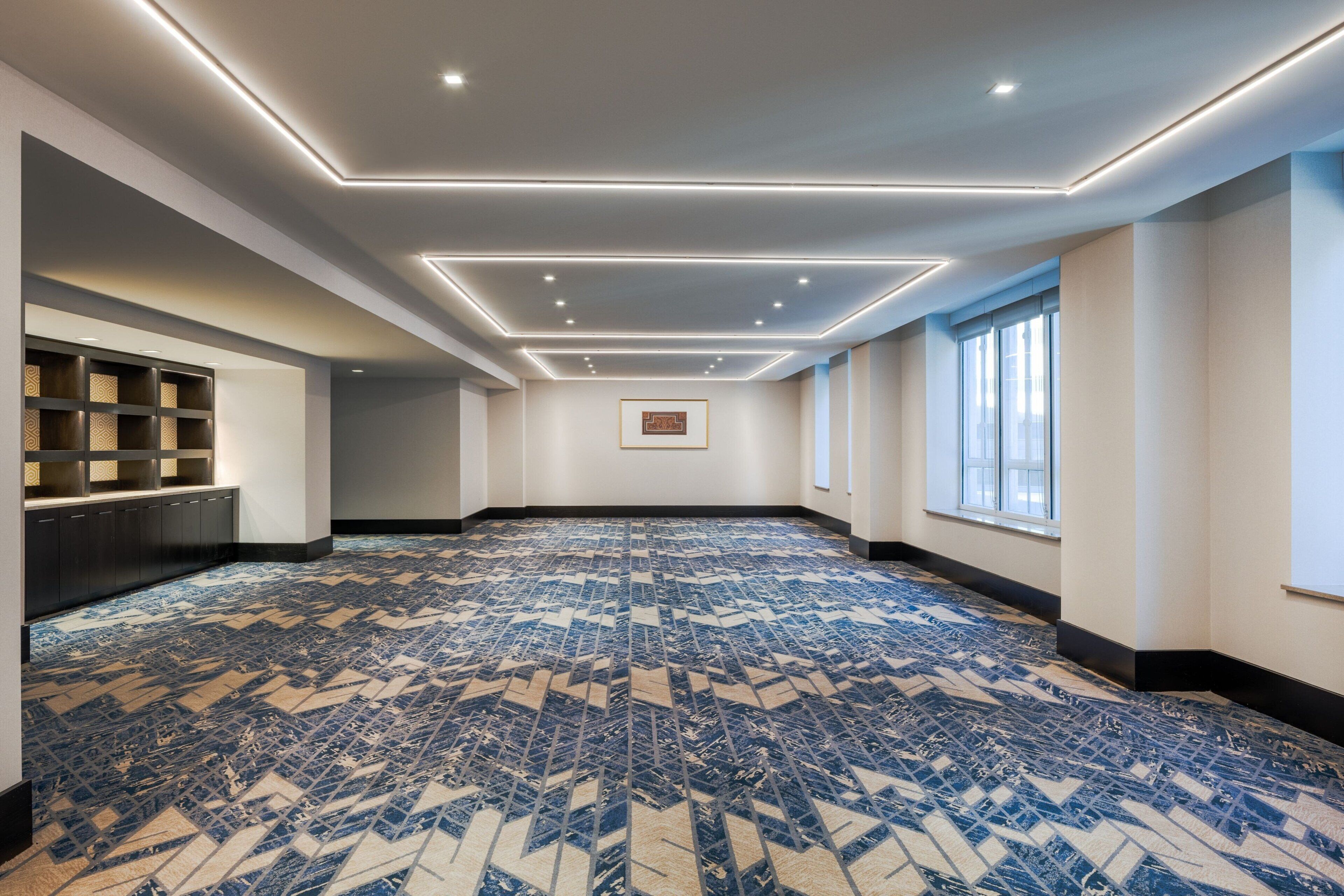 A spacious room with a patterned carpet, recessed ceiling lighting, a framed picture on the wall, and a window on the right side.