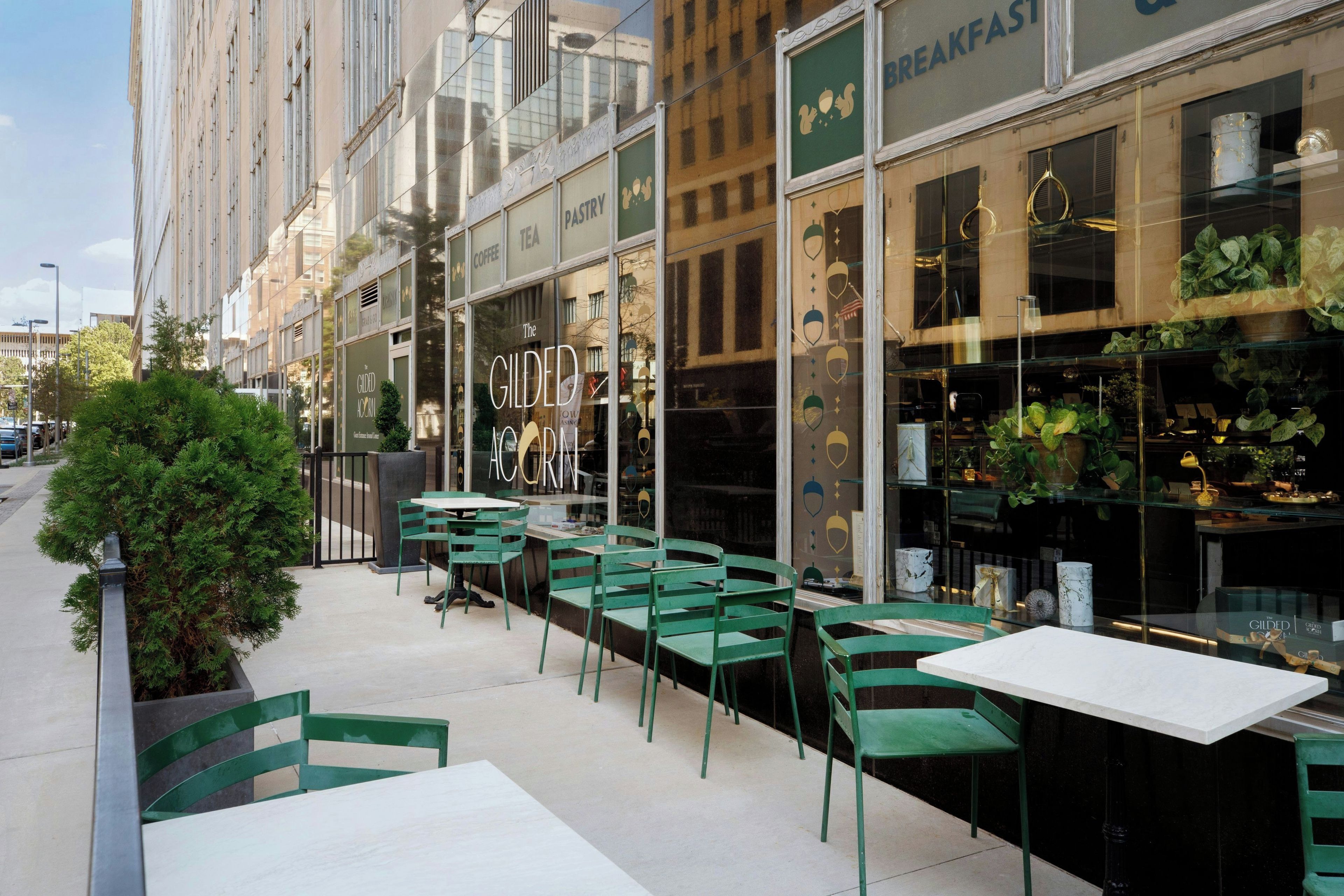 An outdoor seating area with green chairs and tables, a storefront with a sign reading 'BREAKFAST,' and a building with large windows reflecting the sky.