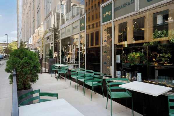 Un área de estar al aire libre con sillas y mesas verdes, un escaparate con un letrero que dice 'BREAKFAST' y un edificio con grandes ventanales que reflejan el cielo.