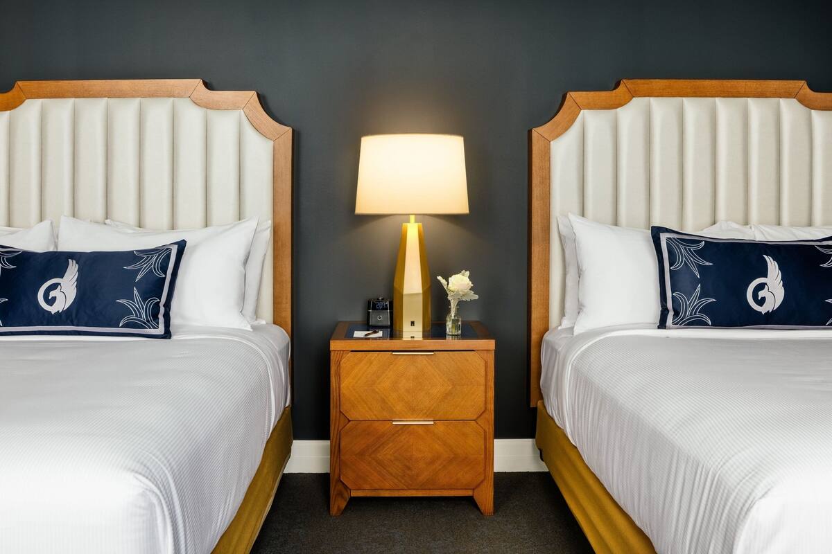 A hotel room with two beds, each with a wooden headboard, a bedside table with a lamp, and matching pillows.