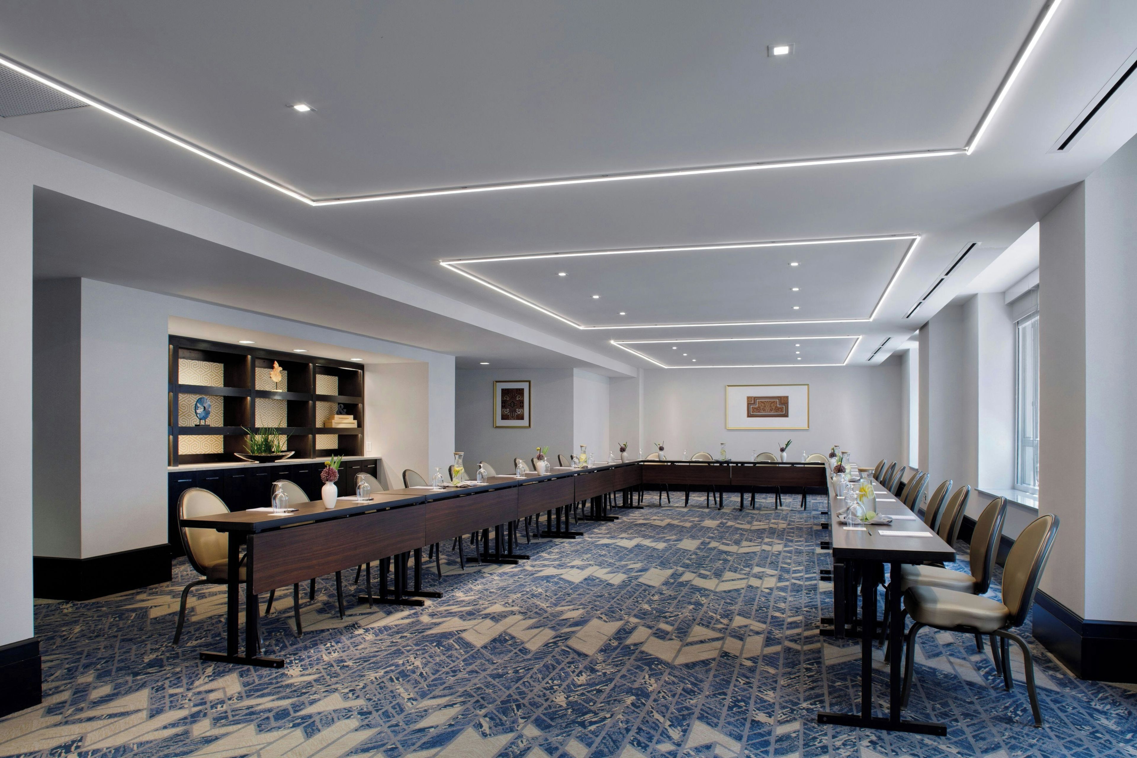 Een vergaderzaal met een lange tafel, stoelen, een kamerbreed tapijt en een boekenkast met decoratieve objecten.