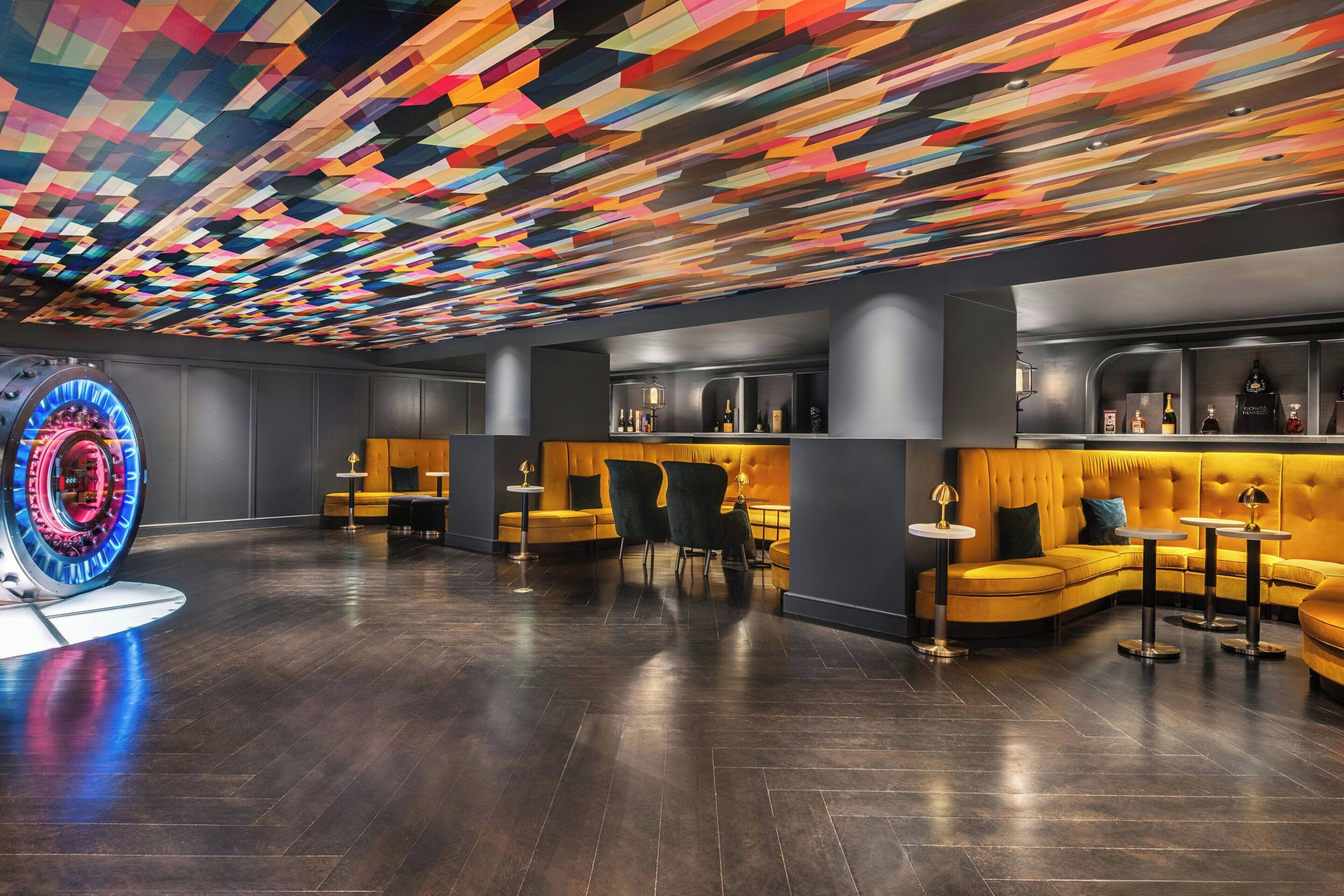 A modern lobby with a colorful geometric ceiling, dark wooden flooring, and a bar area with seating.