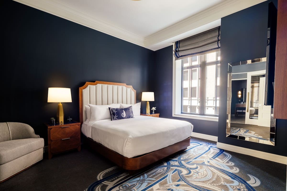 A hotel room with a large bed, two bedside tables with lamps, a sofa, a patterned carpet, and a large mirror.