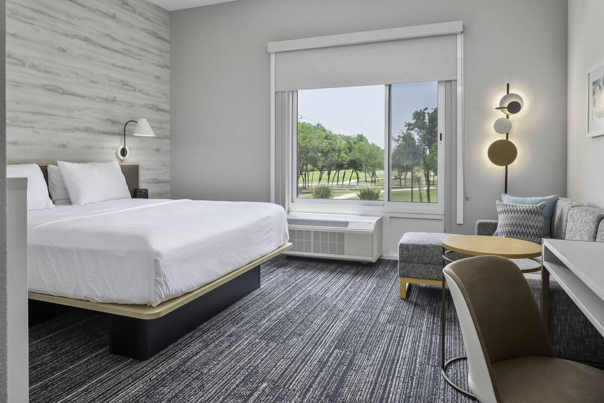 Une chambre d’hôtel équipée d’un lit, d’un bureau, d’une chaise, d’une lampe et offrant une vue sur un parc grâce à une grande fenêtre.