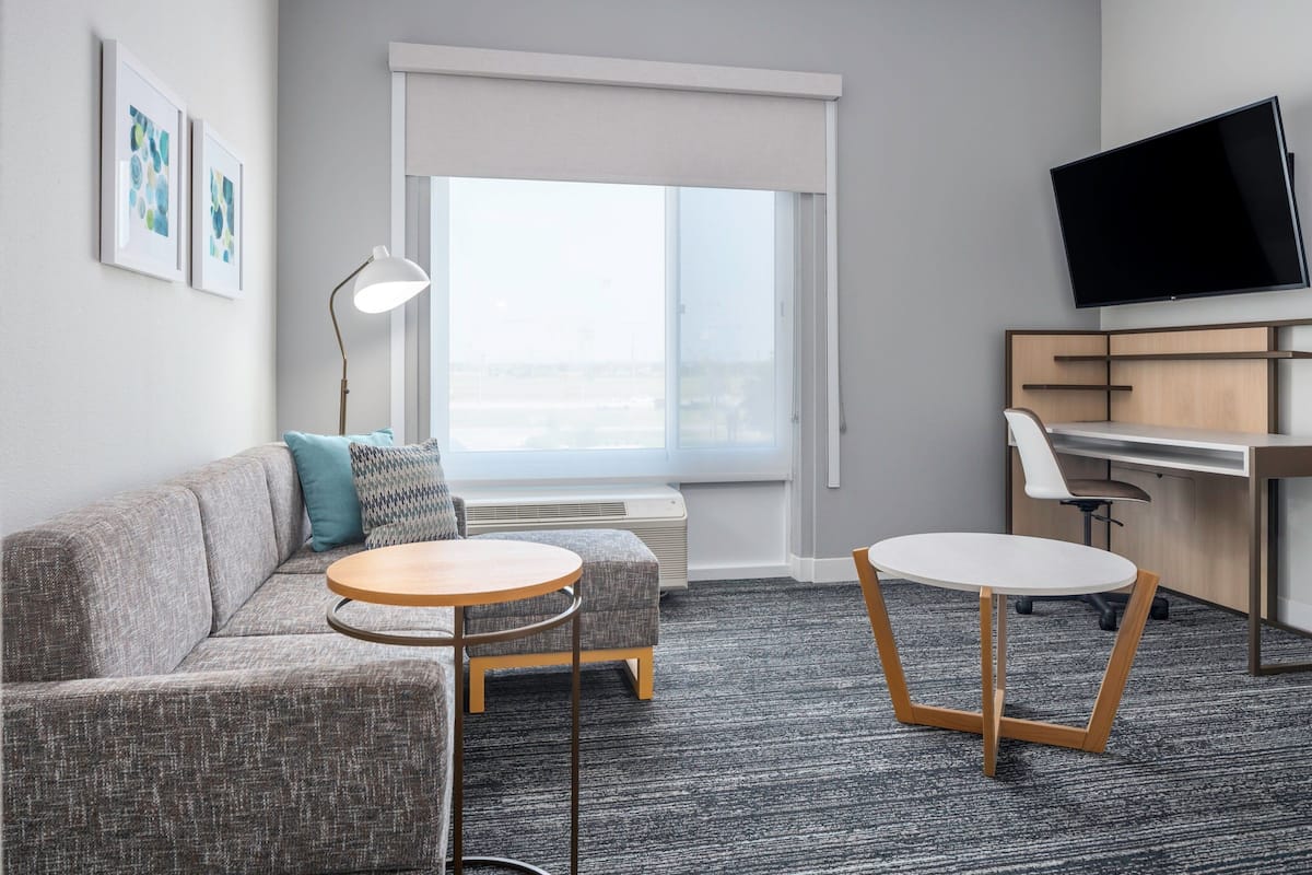 Une chambre d’hôtel moderne avec un canapé gris, deux petites tables rondes, un bureau et un téléviseur à écran plat.