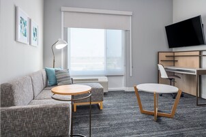 A modern hotel room with a grey sofa, two round tables, a desk, a TV, and a window with blinds.