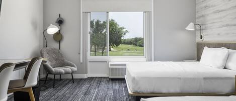 Habitación de hotel con cama, una silla, un escritorio, una lámpara y ventana con vista a un campo de golf.