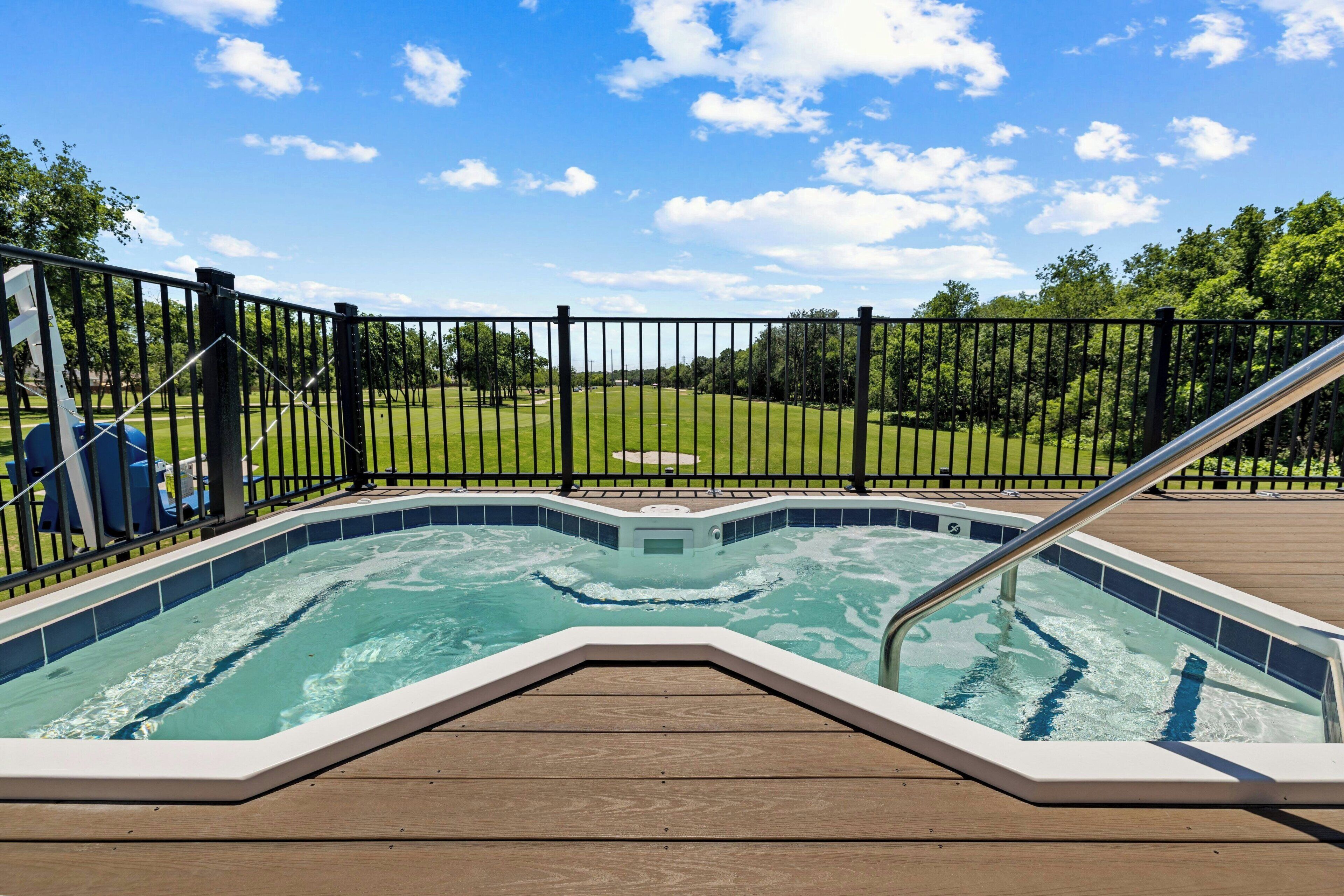 A hot tub on a wooden deck with a metal railing, surrounded by a fence and a grassy area with trees.