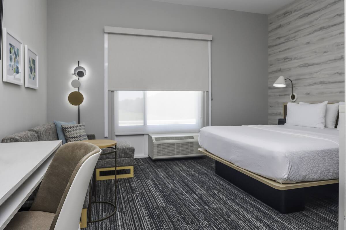 Ein modernes Hotelzimmer mit einem Bett, einem Schreibtisch, einem Sessel, einem kleinen Tisch und einer Wand-Klimaanlage.